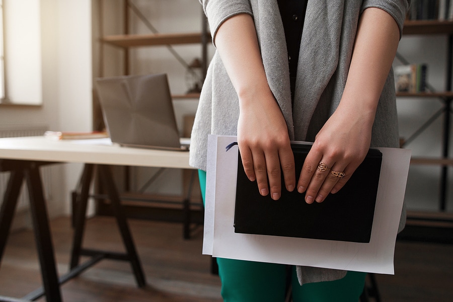 Close-up of a professional woman standing in an office, holding a notebook and documents in her hands. The background shows a desk with a laptop and shelves, creating a modern workplace environment.
